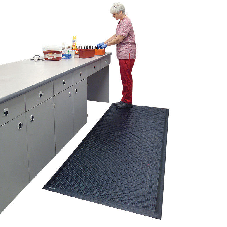 A person stands on a Cushion Station Anti-Fatigue Mat in front of a counter, working with utensils on the surface.