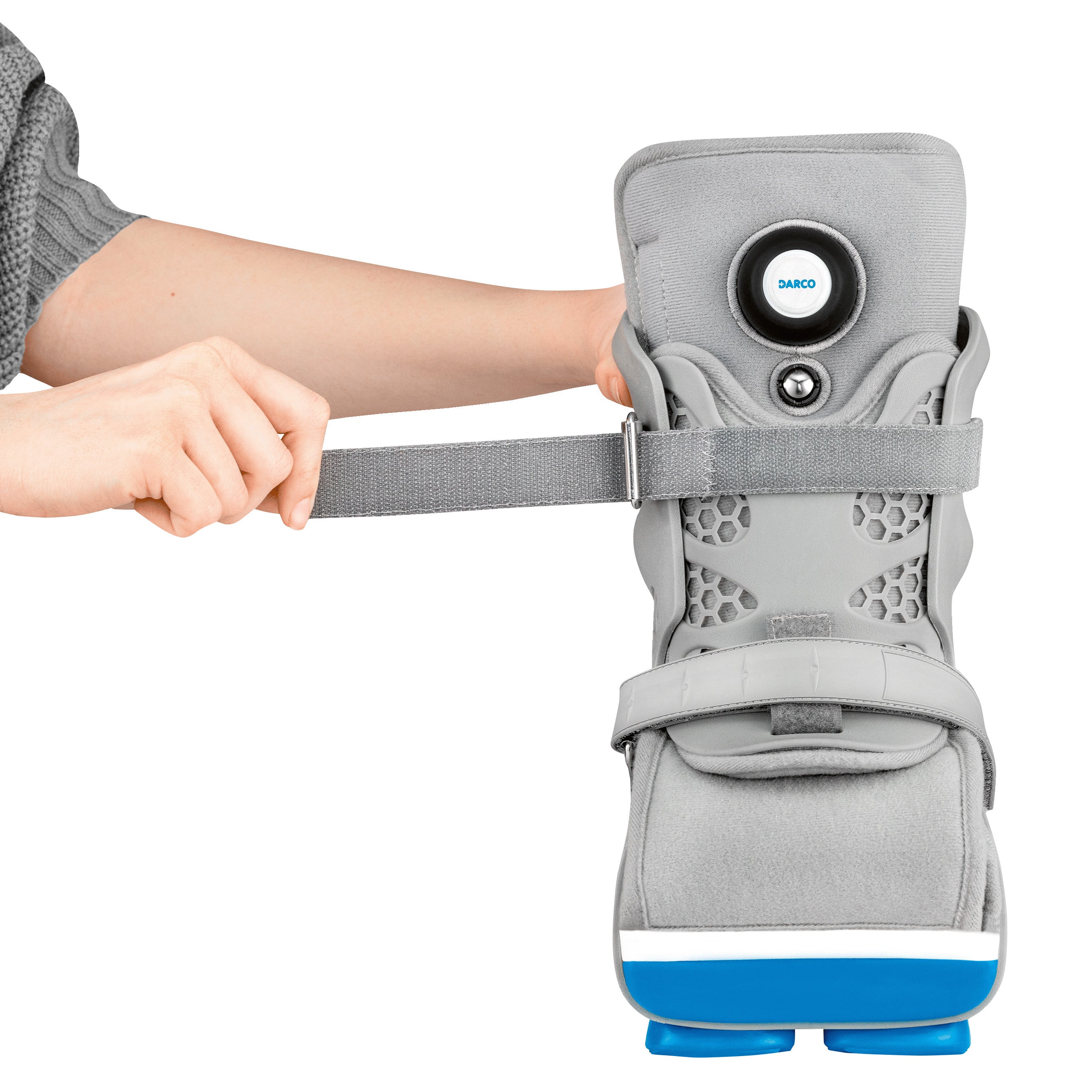 A person is adjusting the strap of a DARCO Body Armor PFA Walker, which is a gray orthopedic boot with blue accents.
