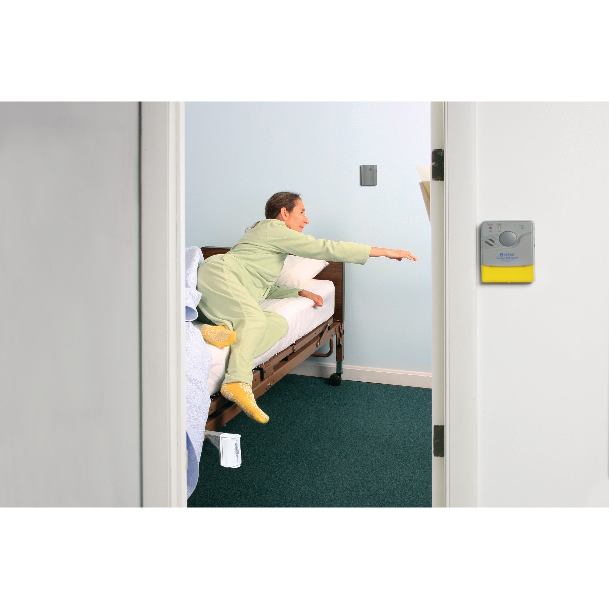 A person reaches from a bed, activating the AliMed Motion Detection Local Alarm with Bed Rail Clamp, mounted on a wall nearby.