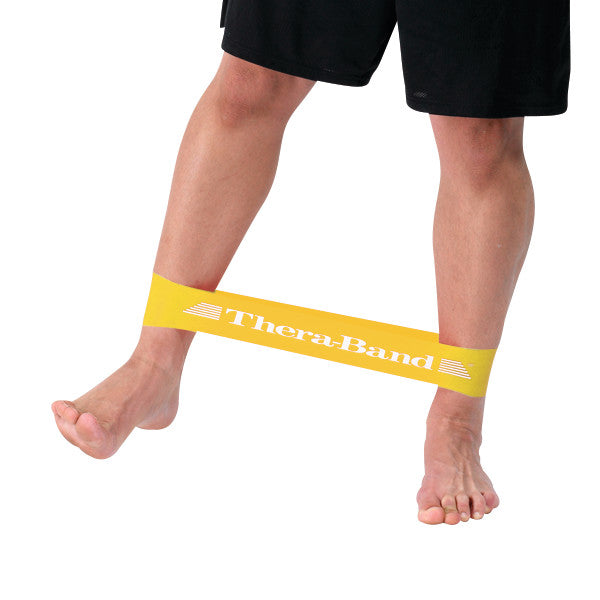 A person demonstrates the THERABAND Resistance Band Loop around their ankles. The band is yellow and branded "Thera-Band."