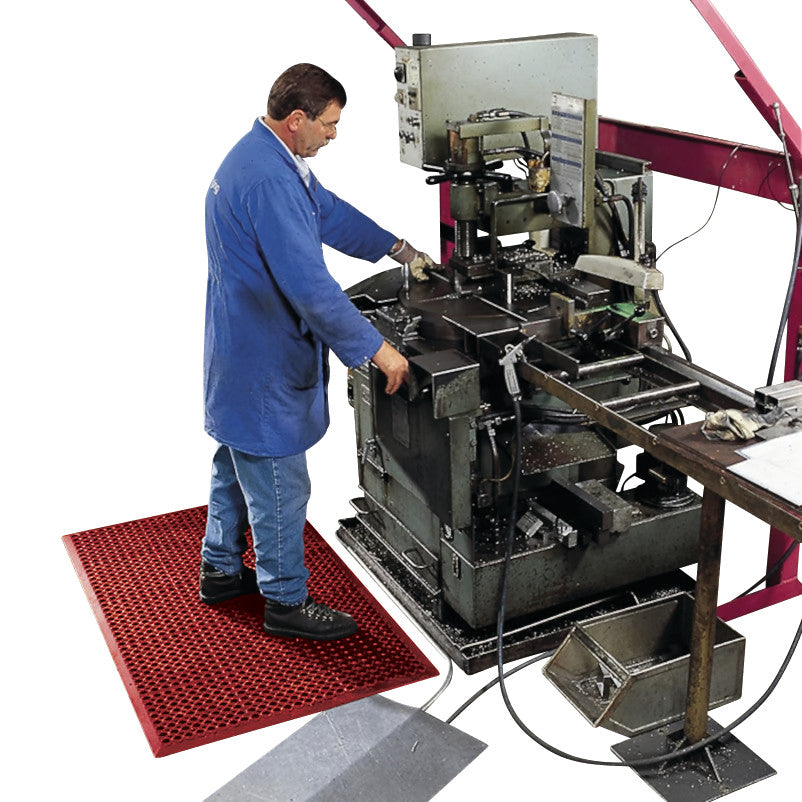 A person stands on a Sanitop Drainage Anti-Fatigue Mat next to machinery. The mat is red with a grid pattern, providing comfort.