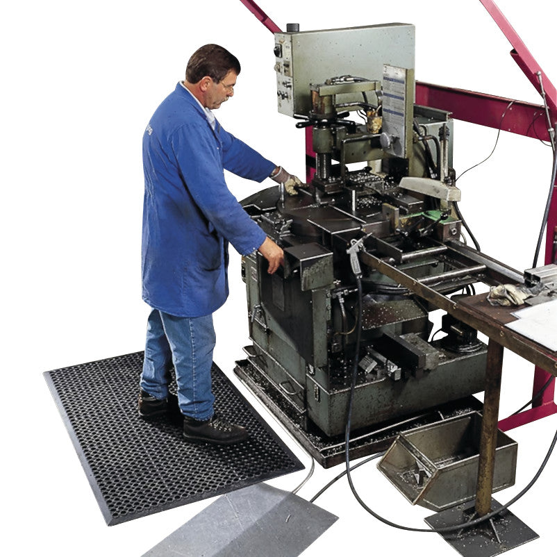 A person stands on a Sanitop Drainage Anti-Fatigue Mat while operating industrial machinery. The mat features a textured surface.
