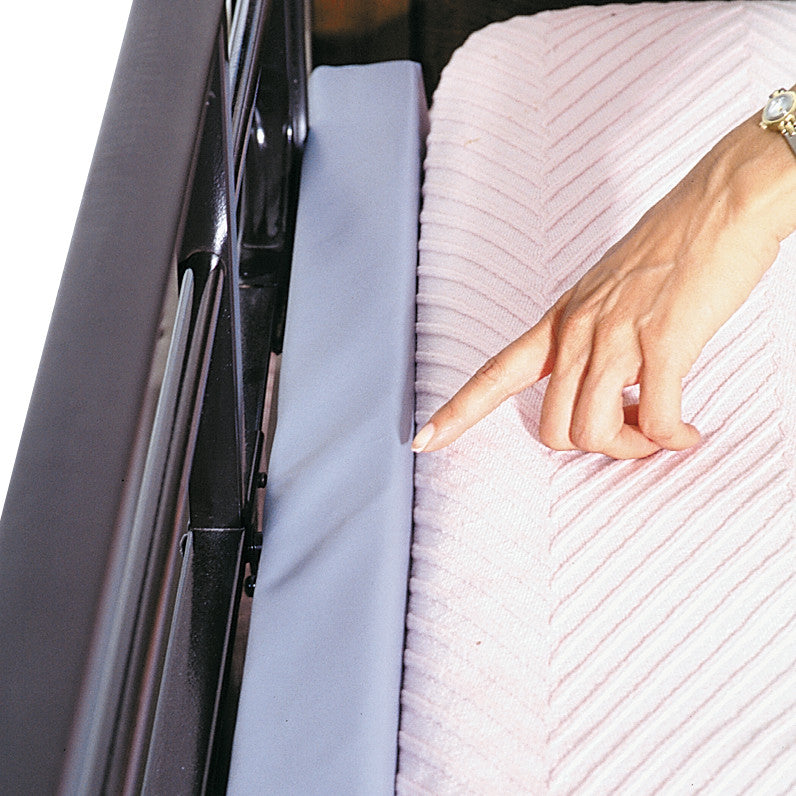 A hand touches the AliMed Bed Stuffer Safety Bolster, positioned on a bed with a pink textured blanket, next to a black bed rail.