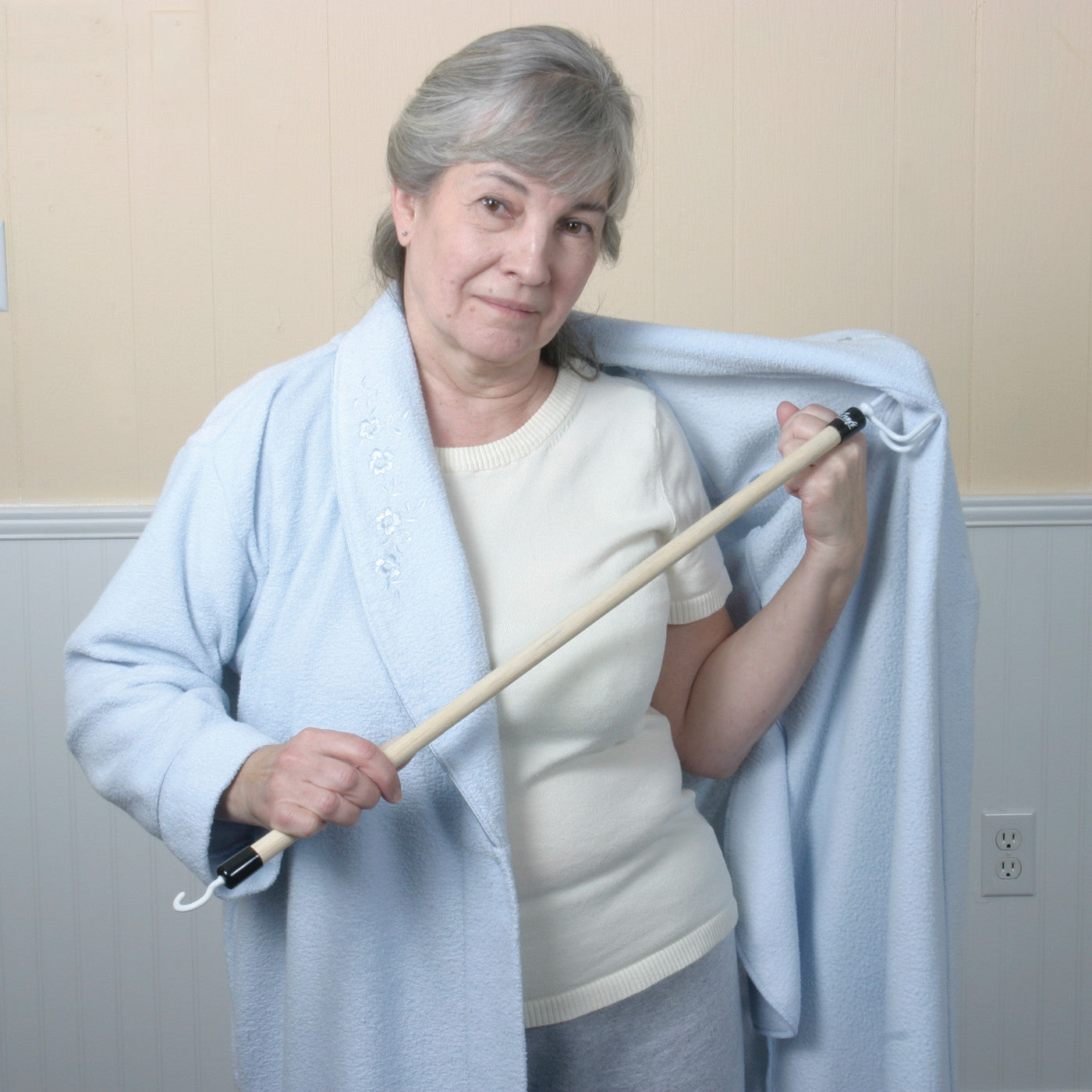 A woman uses the Big Hook Dressing Stick to put on a light blue robe. The stick has a wooden handle and a large hook.