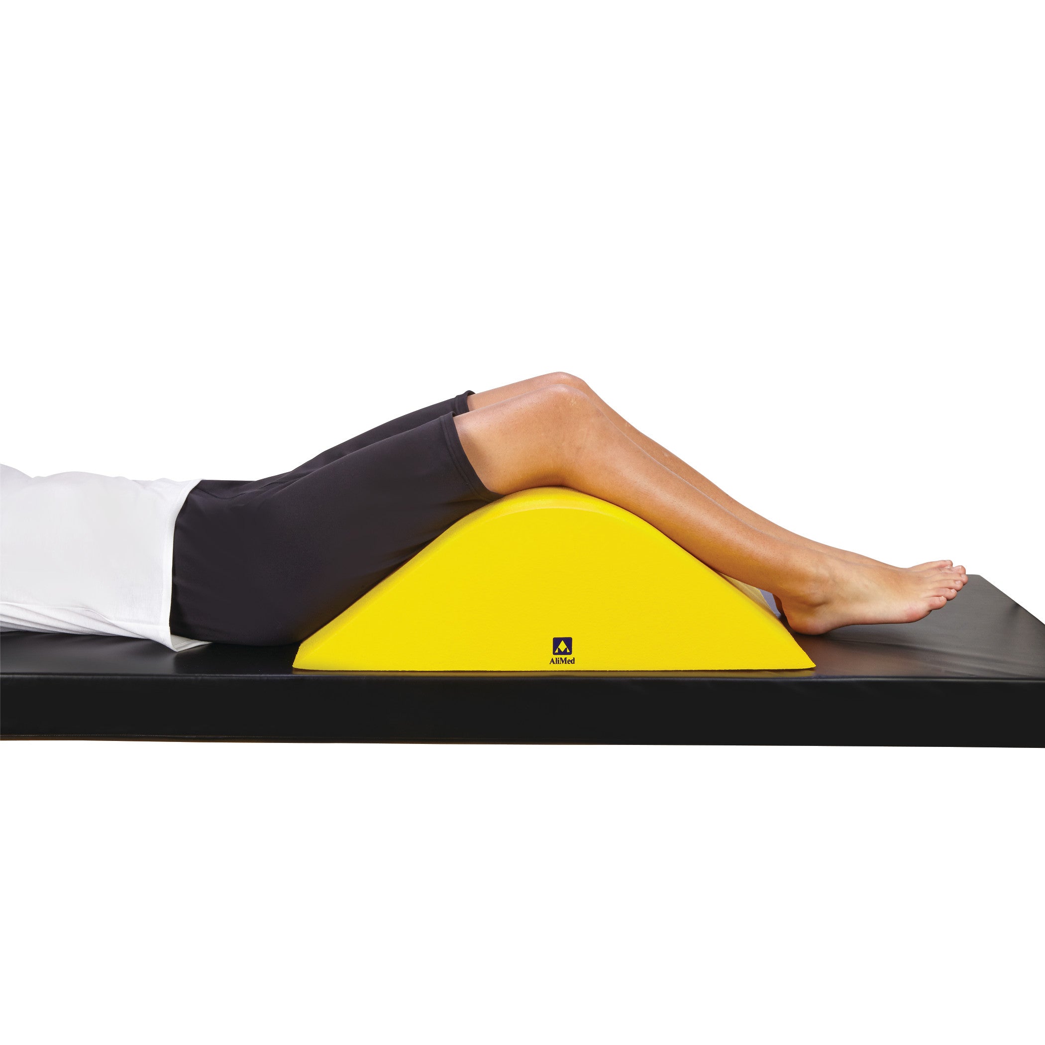 A person rests their legs on the yellow AliMed Protecta-Coat Deluxe Knee Bolster, placed on a black surface for support.