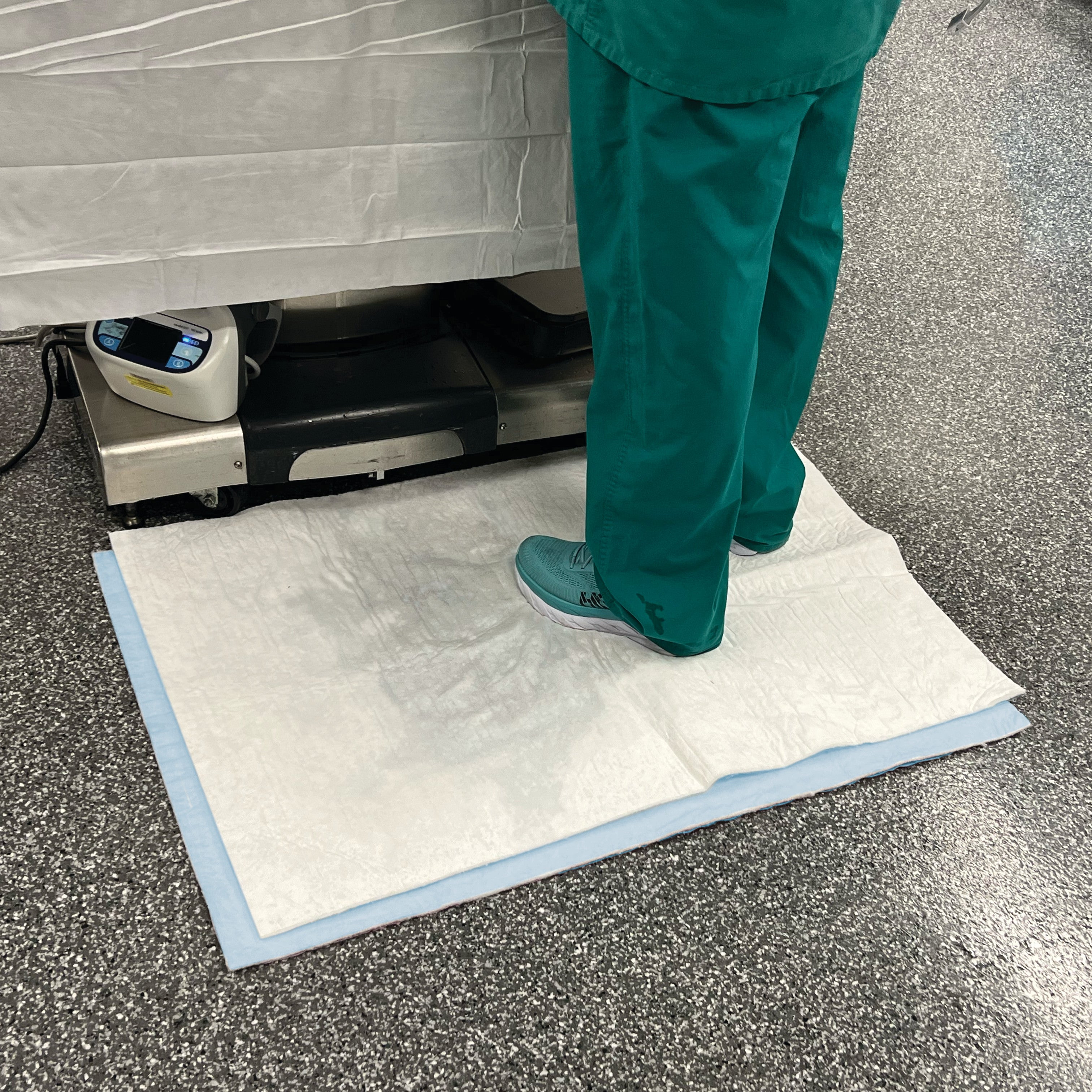 A person stands on a SpilfyterMED White Absorbent Mat for Emergent Use, wearing teal scrubs and shoes, on a speckled floor.