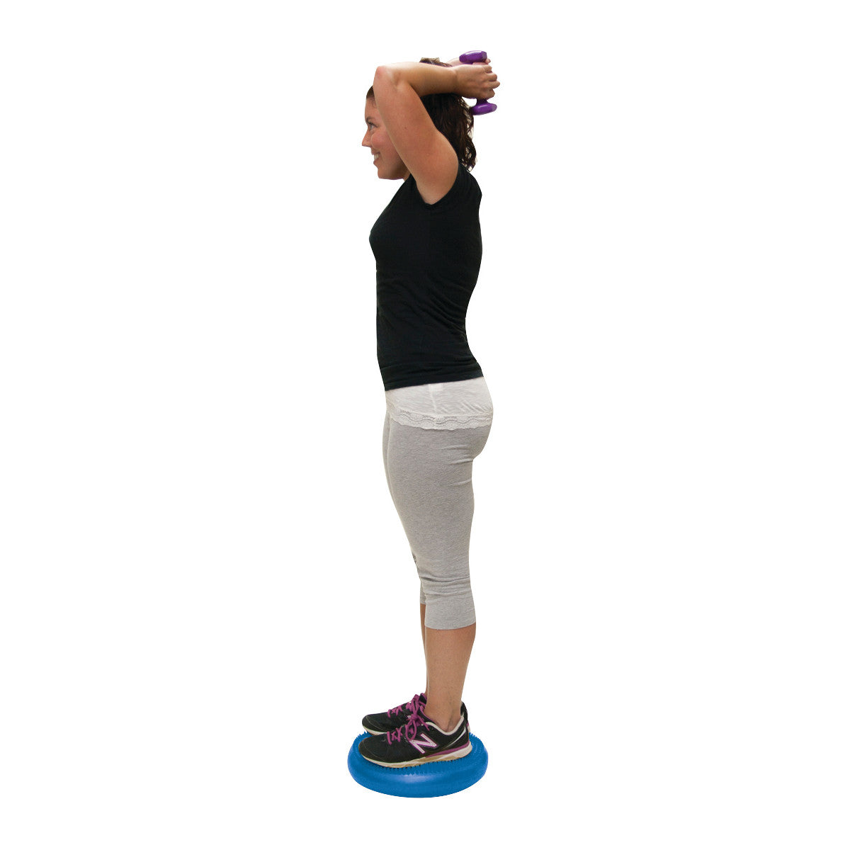 A woman exercises by standing on a CanDo Vestibular Disc, holding a small dumbbell above her head, wearing athletic clothing.