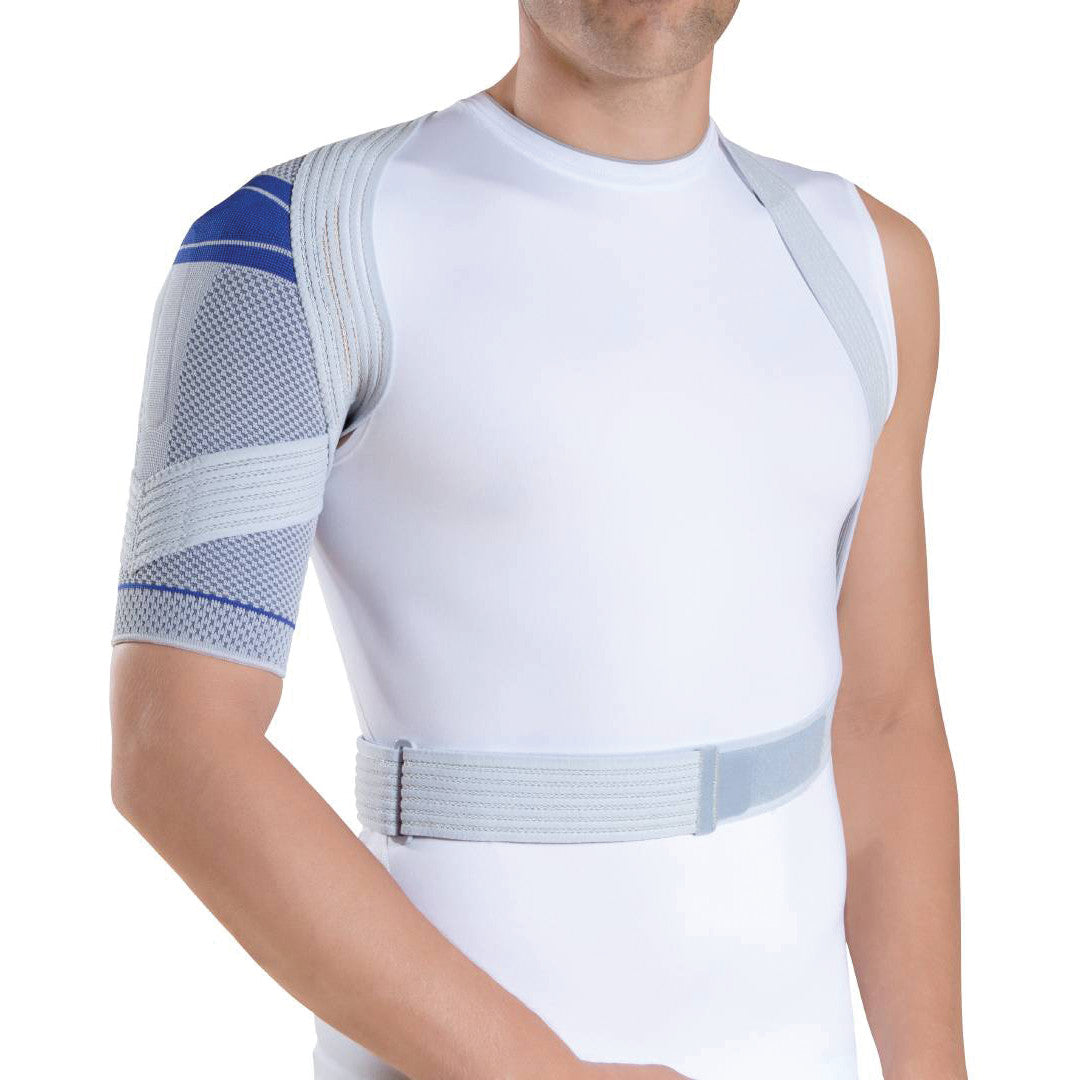 A man wearing a white tank top is fitted with a Bauerfeind OmoTrain Active Shoulder Support, featuring adjustable straps and a knitted sleeve.