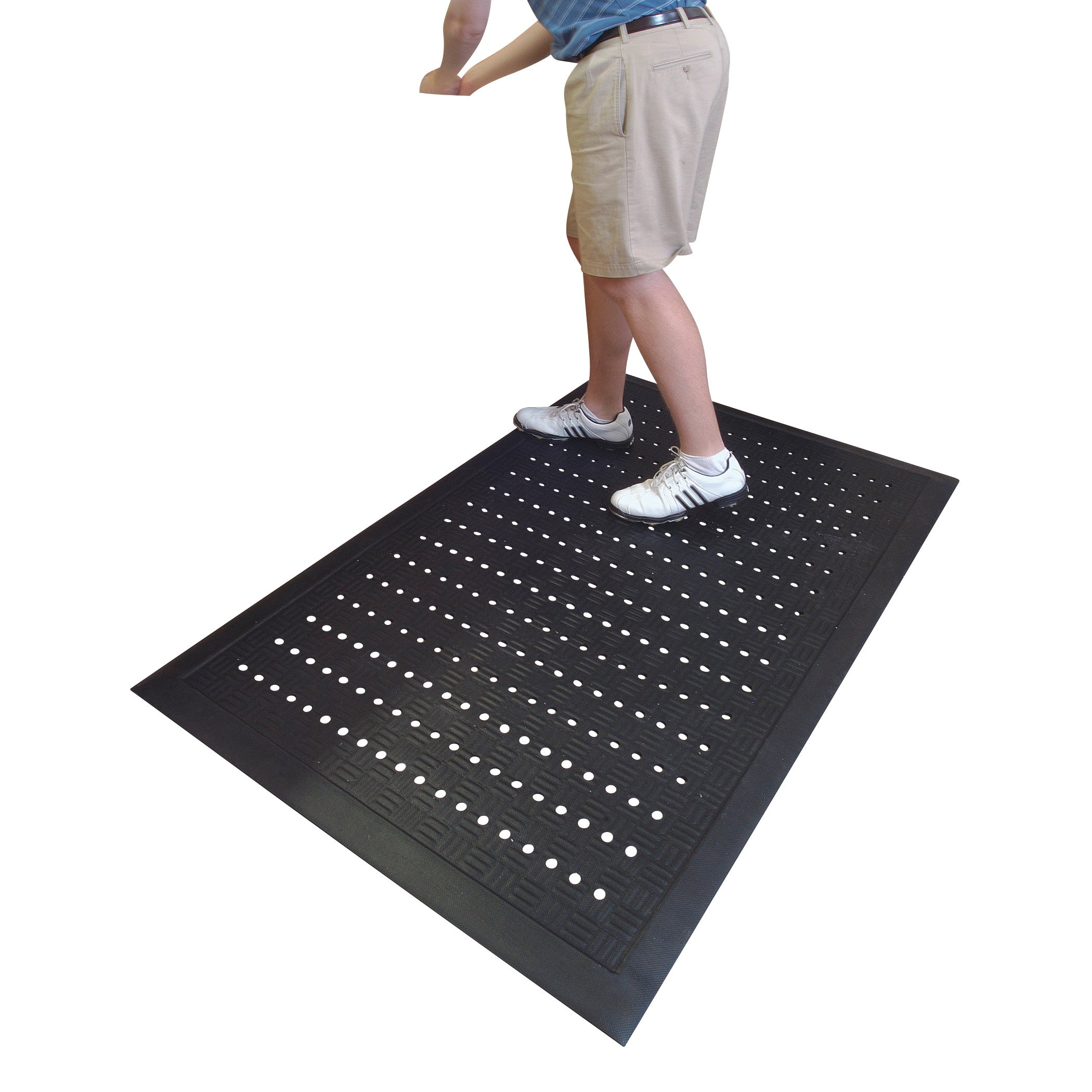 Person standing on a black Cushion Station Anti-Fatigue Mat with small holes and textured edges, wearing shorts and sneakers.