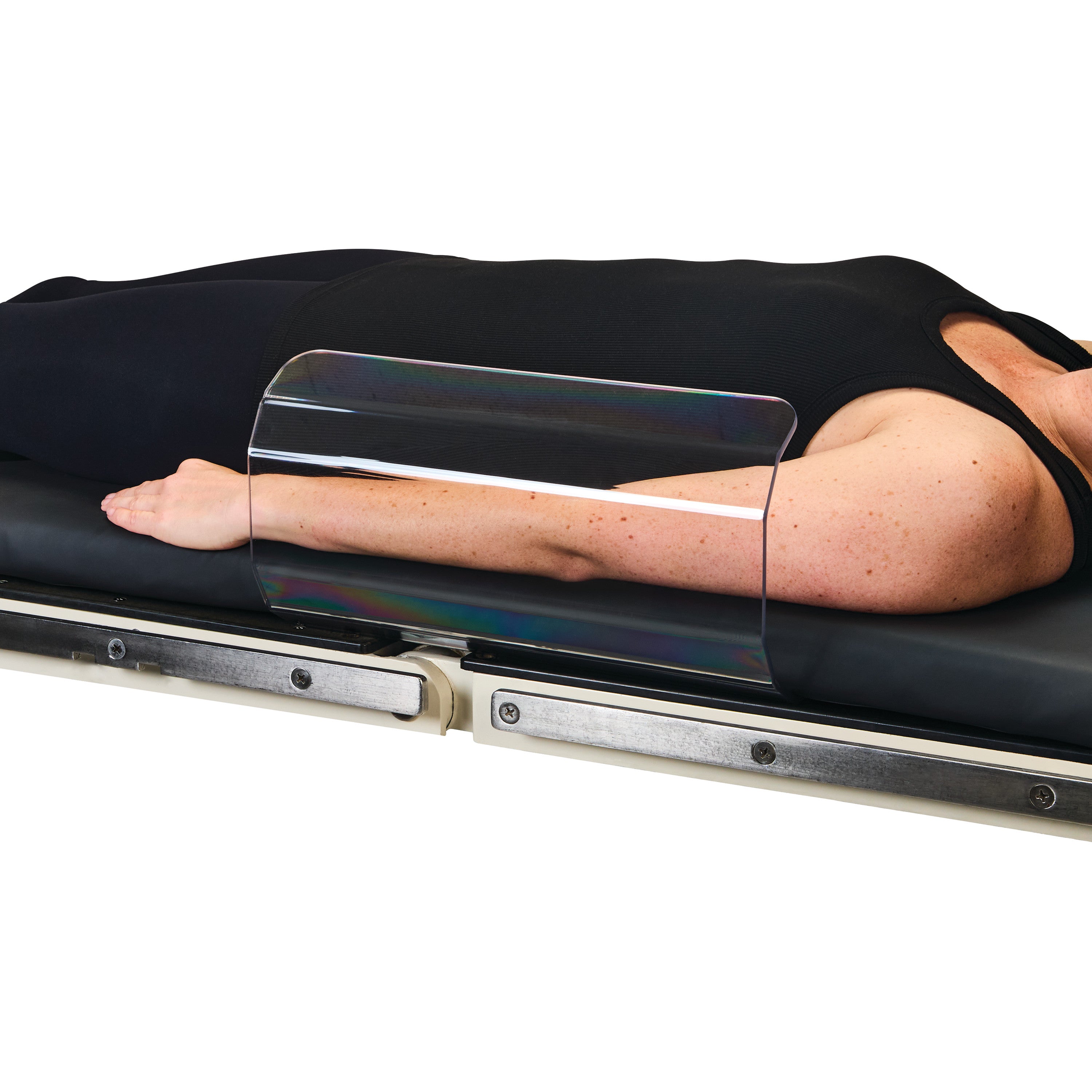 A person is lying on a table with their arm secured under the AliMed Curved Toboggan Arm/Leg Guard, a transparent protective barrier.