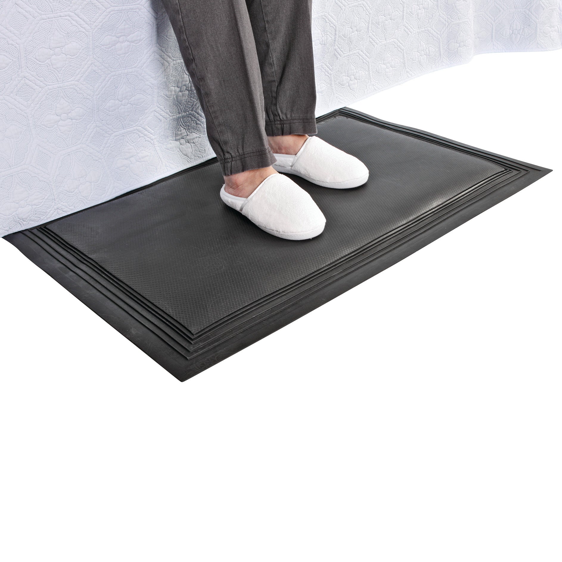 A person wearing white slippers stands on an AliMed Nonslip Exit Alarm Mat beside a bed with a white quilted cover.