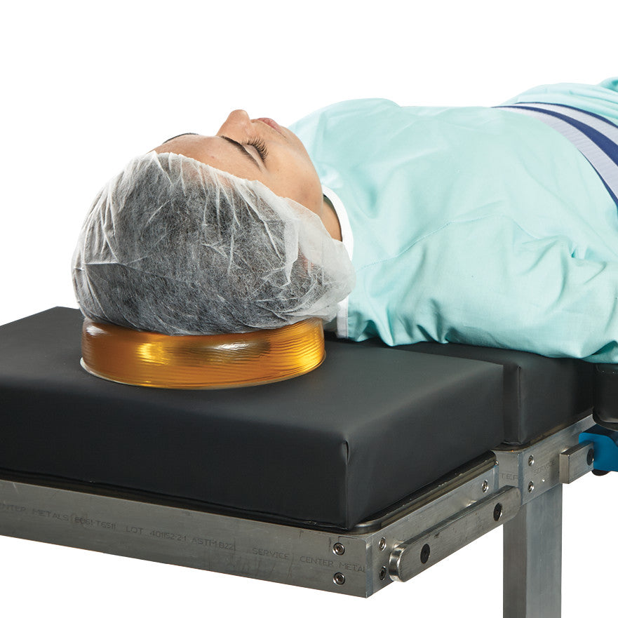 A person lies with their head on an AliMed AliGel Head Donut, placed on a medical table. They wear a hairnet and hospital gown.