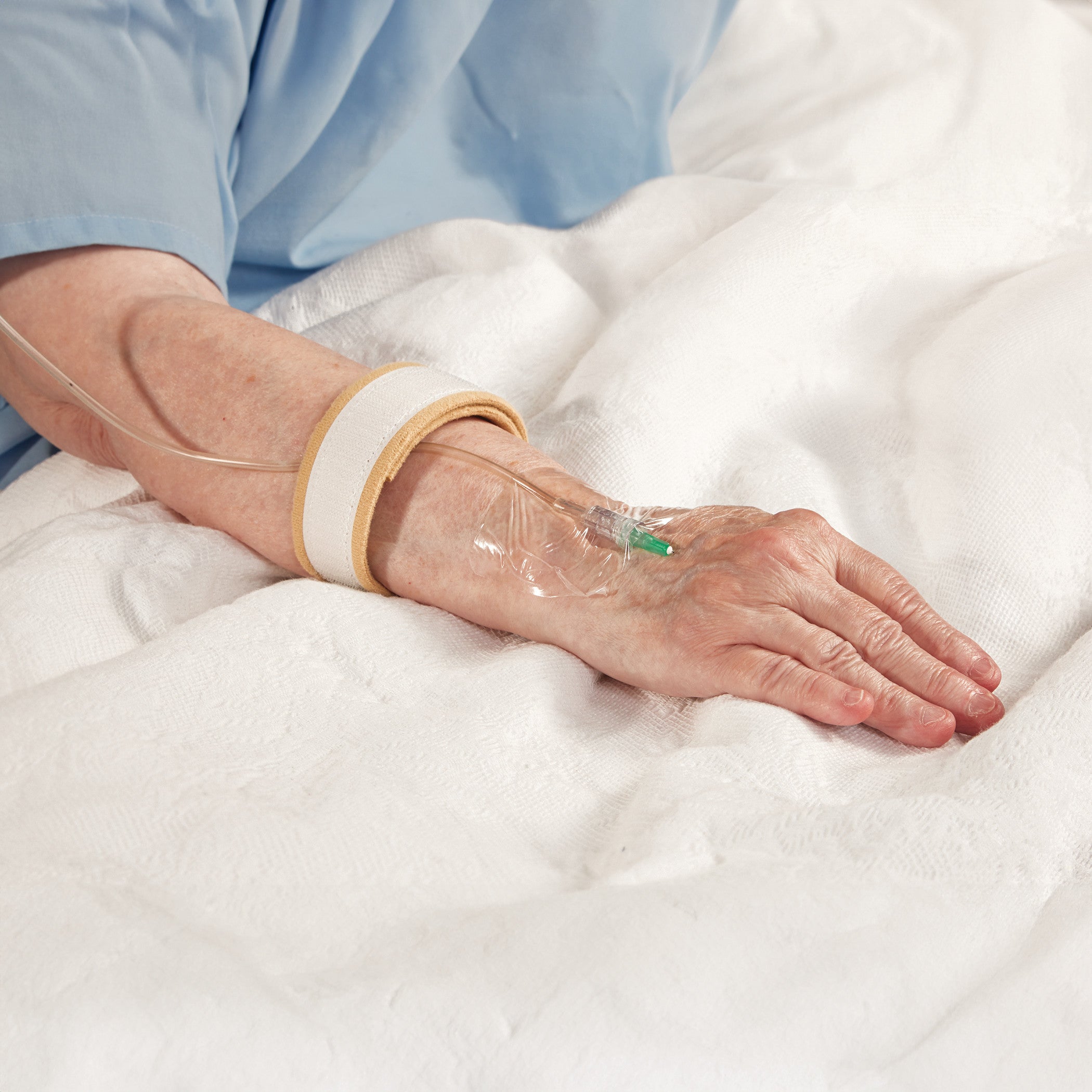 An elderly patient with an IV is resting. The AliMed AliStrap Soft Positioning Strap secures their arm on a white bed.